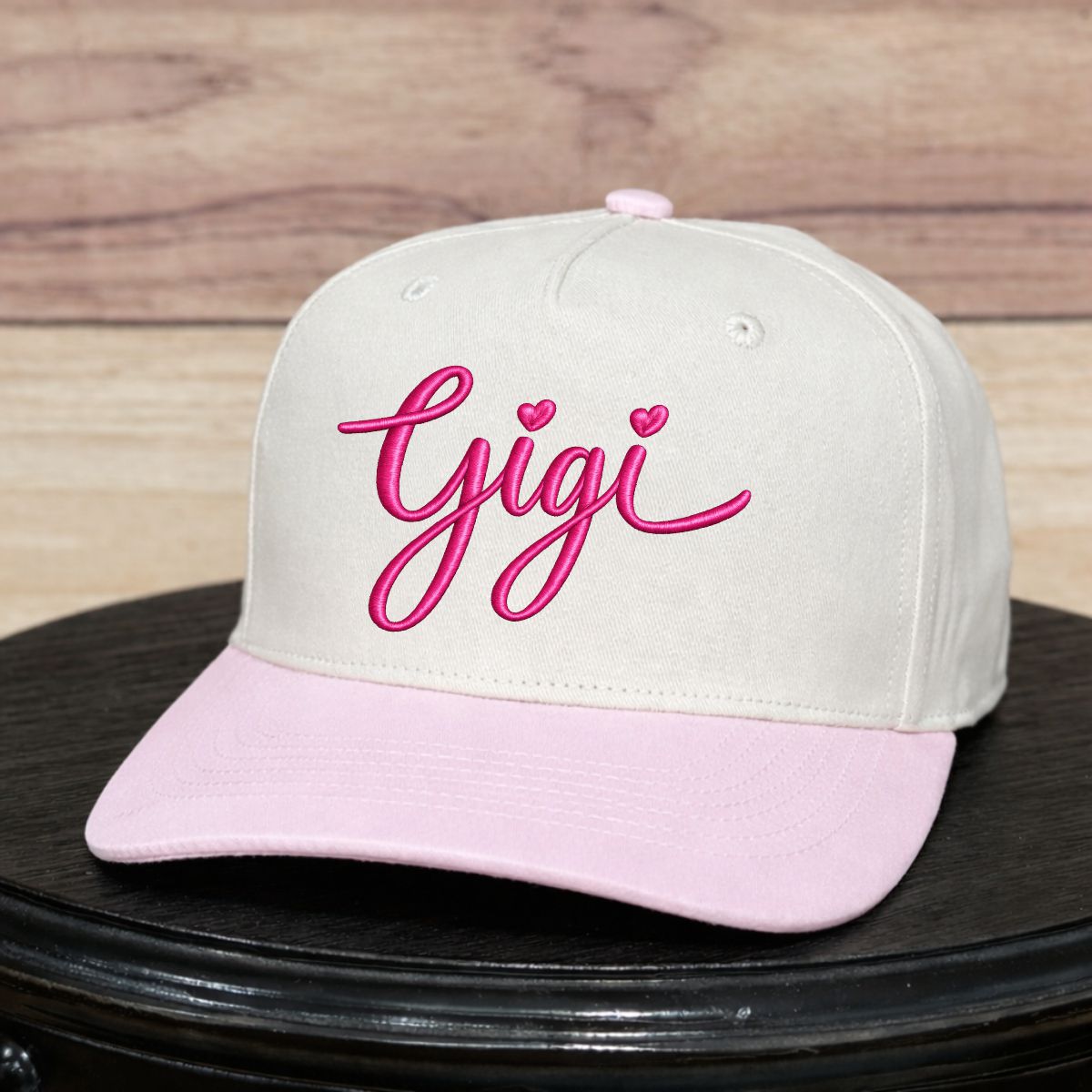 Beige cap with pink brim and 'gigi' text on a wooden surface