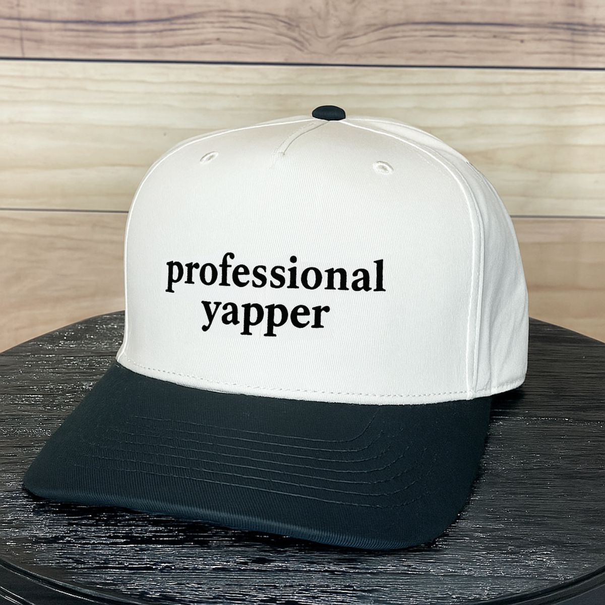 White and black cap with 'professional yapper' text on a wooden surface
