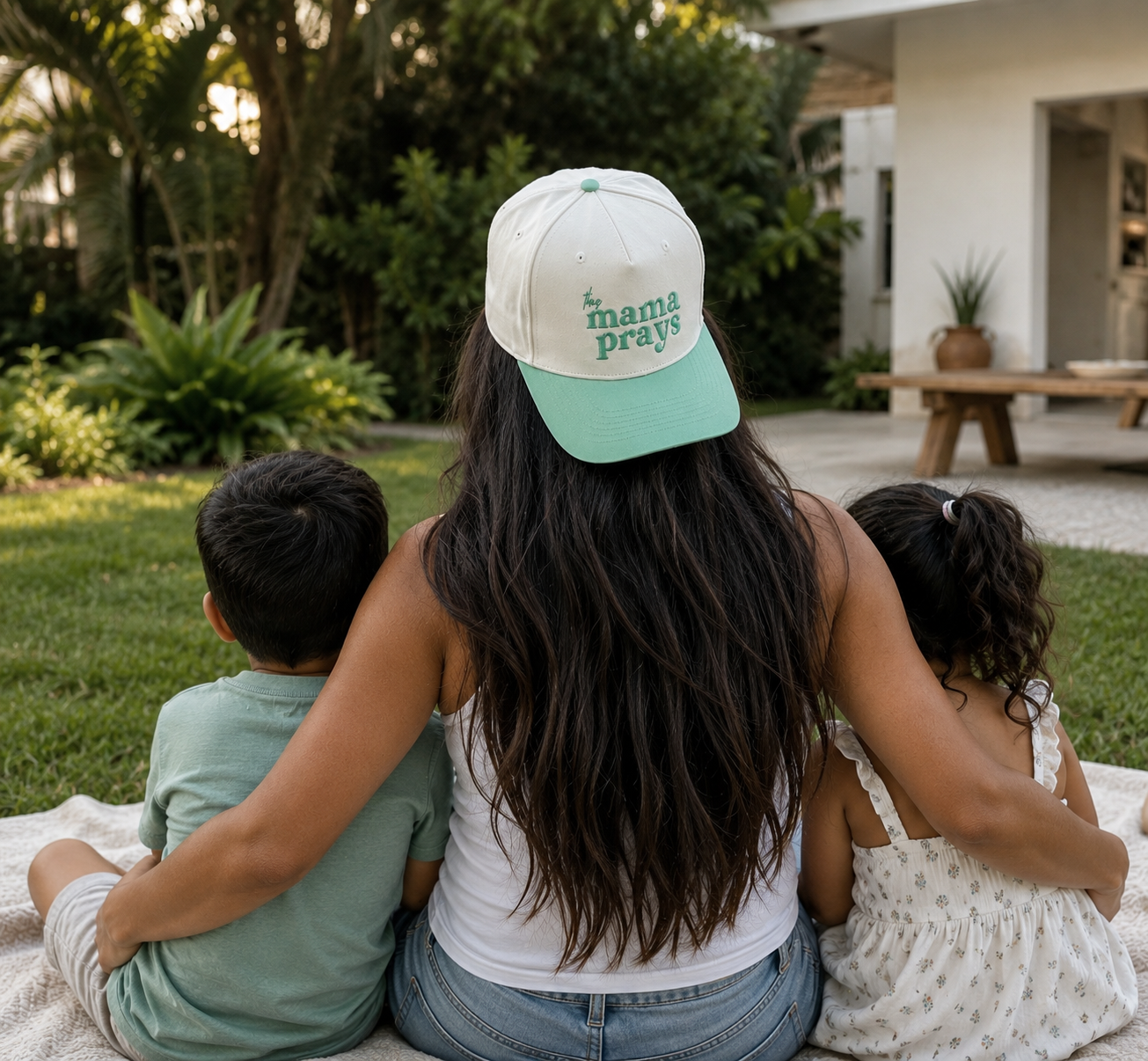 A woman sitting outdoors on a blanket with two children, viewed from behind. She is wearing a white and mint green baseball cap with the text 'this mama prays' printed on the back.