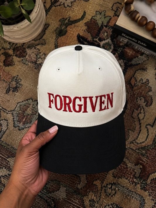 A hand holds a two-tone baseball cap with an off-white crown and a black curved brim. The front of the cap features the word 'FORGIVEN' embroidered in red block letters.