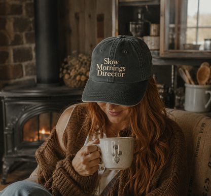 A woman in a cozy living room setting wearing a dark gray baseball cap with 'Slow Mornings Director' embroidered in gold, while holding a ceramic mug.