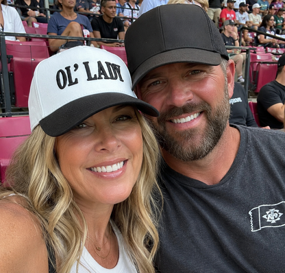 A smiling couple posing together in a stadium crowd. The woman wears a white and black trucker hat with 'OL' LADY' printed on it, and the man wears a black mesh-back cap and a dark gray t-shirt with a small white flag logo.