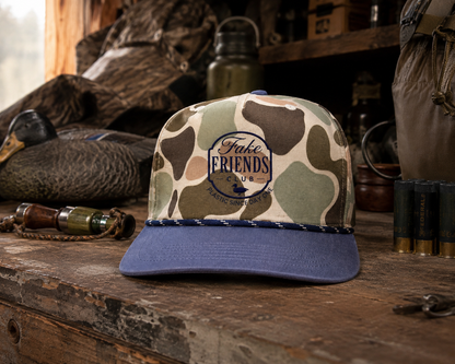 Fake Friends Duck Club embroidered duck hunting hat featuring a camouflage crown and navy bill, styled with duck decoys, shotgun shells, and hunting gear in a rustic duck blind.