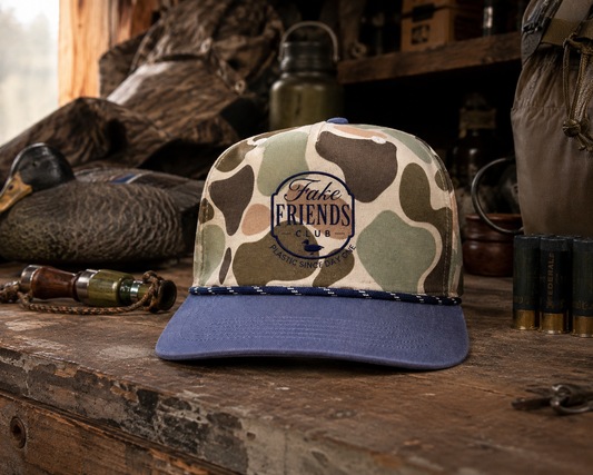 Fake Friends Duck Club embroidered duck hunting hat featuring a camouflage crown and navy bill, styled with duck decoys, shotgun shells, and hunting gear in a rustic duck blind.