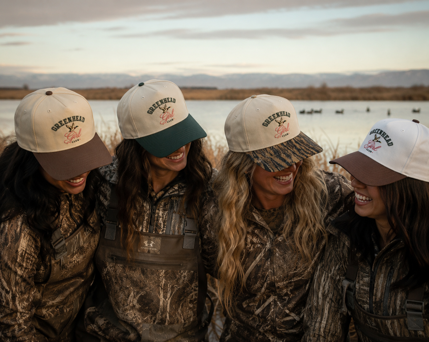 Four women in camouflage hunting jackets and waders stand outdoors by a lake at sunset, wearing matching trucker hats 'Greenhead' mallard duck hunting hats.