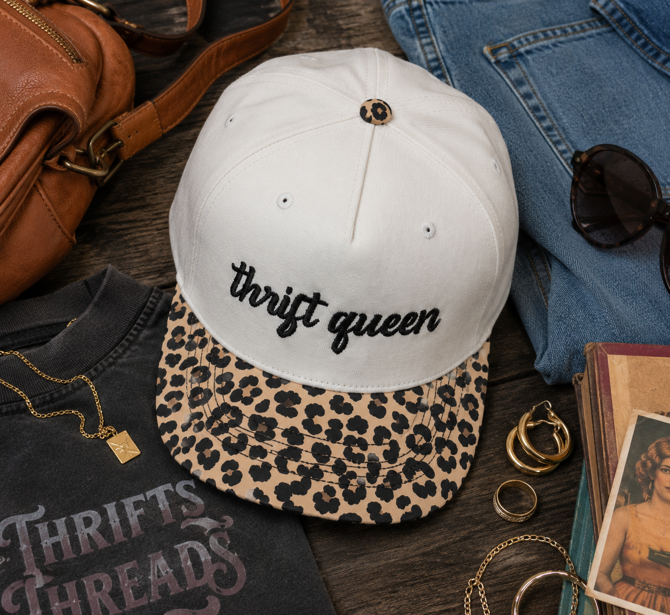 A natural baseball cap with a leopard-print brim and the words 'thrift queen' embroidered in black script on the front. The cap is styled in a flat-lay arrangement with a black graphic t-shirt, blue jeans, sunglasses, a brown leather bag, and gold jewelry on a dark wood surface.