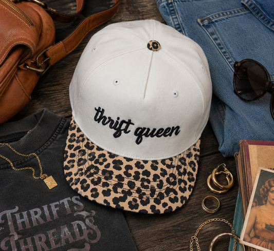 A natural baseball cap with a leopard-print brim and the words 'thrift queen' embroidered in black script on the front. The cap is styled in a flat-lay arrangement with a black graphic t-shirt, blue jeans, sunglasses, a brown leather bag, and gold jewelry on a dark wood surface.