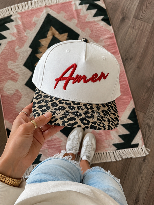 A beige baseball cap with a leopard-print brim and red embroidered 'Amen' script, held by a person wearing white sneakers and light blue denim shorts over a patterned rug.