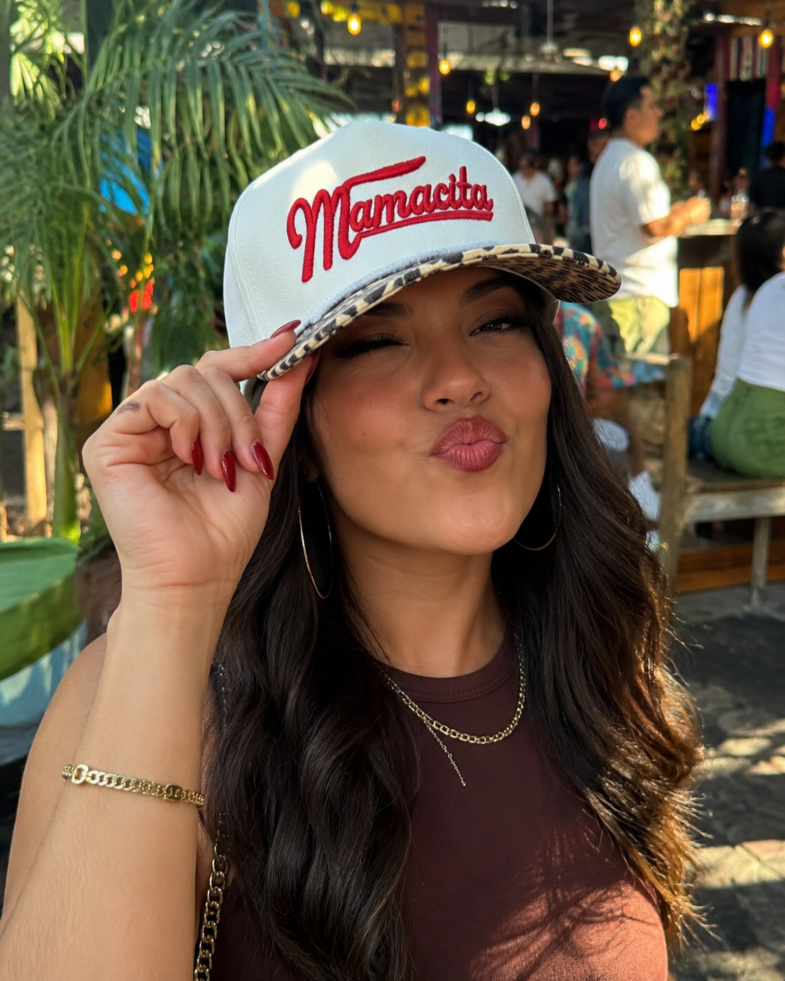 A woman wearing a beige snapback cap with red ‘Mamacita' text and a leopard-print brim, posing outdoors in a sunny, tropical restaurant setting.