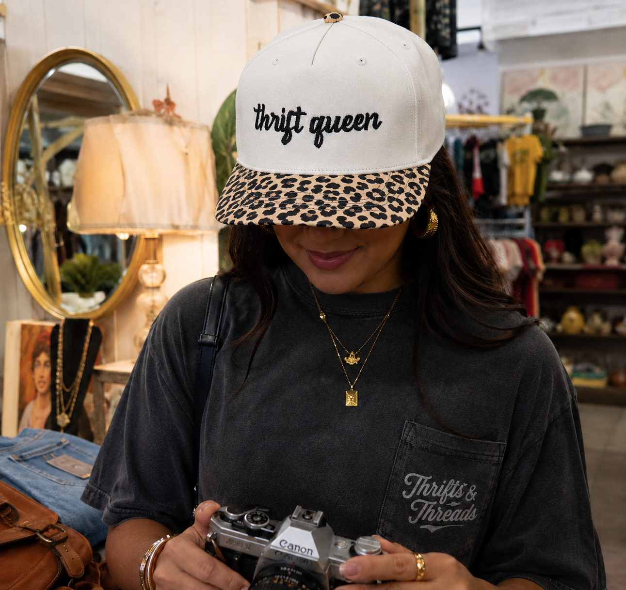 A woman wearing a beige baseball cap with 'thrift queen' embroidered in black on the front and a leopard-print brim, holding a vintage Canon camera in a boutique setting. She is also wearing a dark gray 'Thrift & Threads' t-shirt.