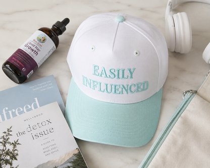 A beige and mint green baseball cap with the words 'EASILY INFLUENCED' embroidered on the front, styled in a flat lay with a bottle of liquid, a magazine, white headphones, and a notebook on a light marble surface.