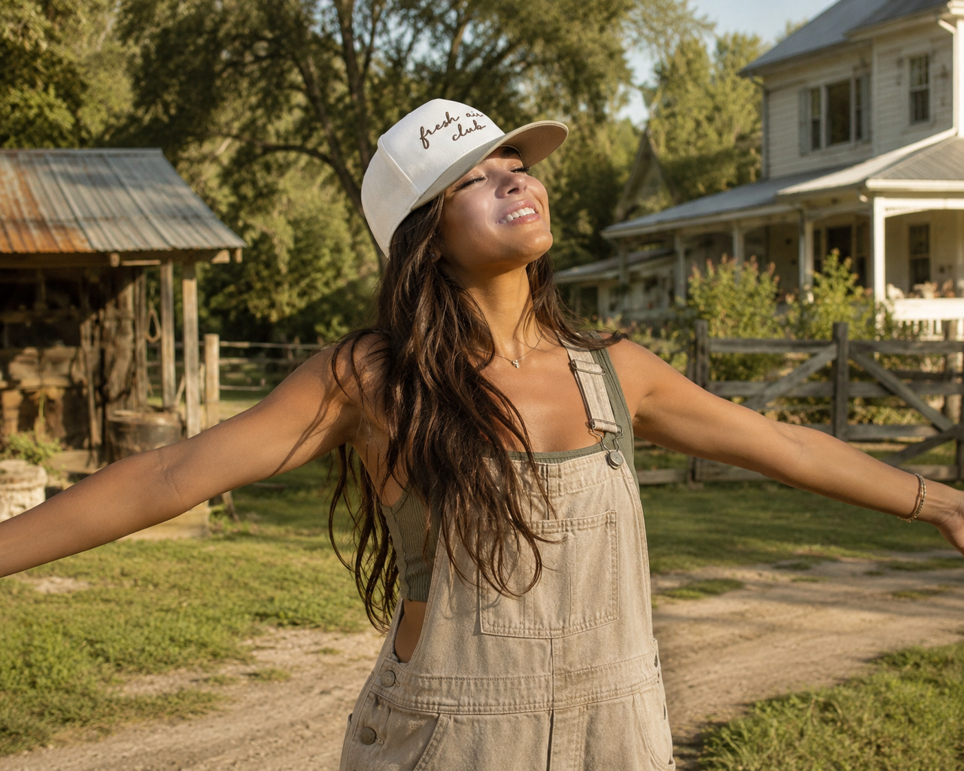 A woman with long dark hair stands outdoors on a farm with her arms outstretched, wearing a natural and khaki cap with brown script ‘Fresh Air Club’ and beige denim overalls.