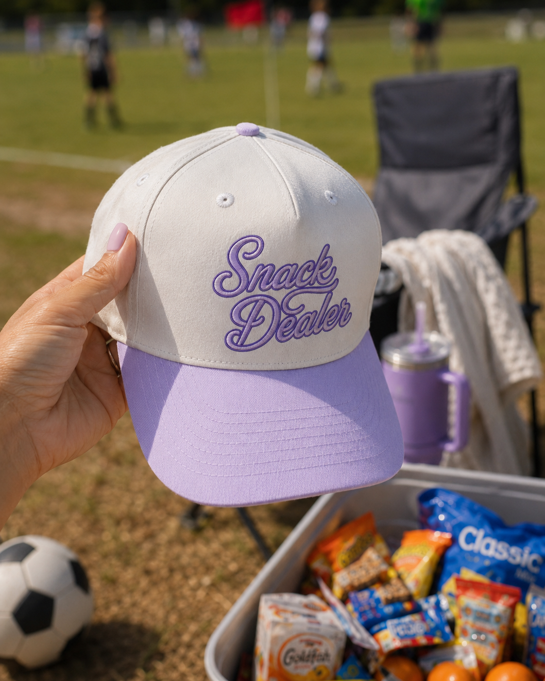 A hand holds a beige and lavender baseball cap with 'Snack Dealer' embroidered in purple script. The background shows a sunny outdoor soccer field with players, a folding chair, a tumbler, and a cooler filled with snacks.