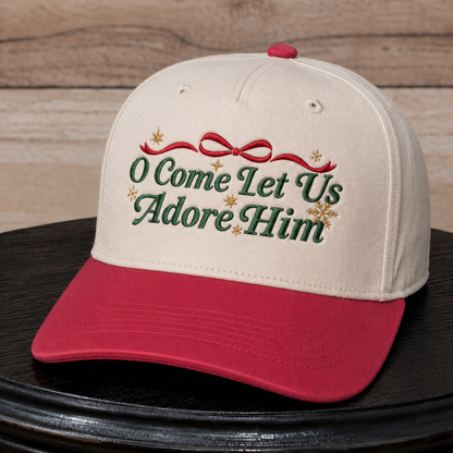 A baseball cap with an off-white crown and a red curved brim, featuring embroidered green text reading 'O Come Let Us Adore Him' with a red bow and gold star details, displayed on a black surface against a wood panel background.