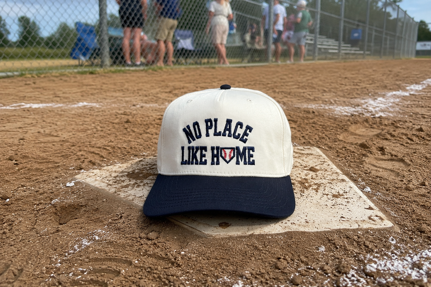 A baseball cap resting on a home plate at a baseball field, with dirt, chain-link fence, and blurred spectators in the background. The cap has an off-white crown with navy blue text reading 'NO PLACE LIKE HOME' and a navy blue brim.