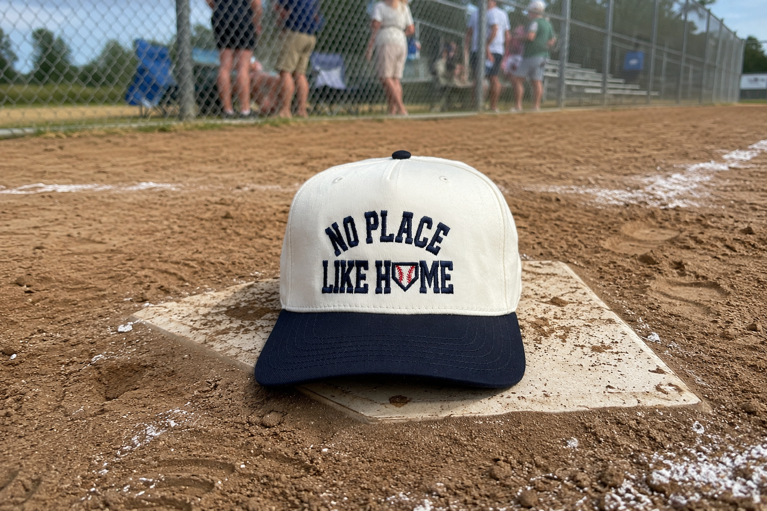 A baseball cap resting on a home plate at a baseball field, with dirt, chain-link fence, and blurred spectators in the background. The cap has an off-white crown with navy blue text reading 'NO PLACE LIKE HOME' and a navy blue brim.