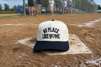 A baseball cap resting on a home plate at a baseball field, with dirt, chain-link fence, and blurred spectators in the background. The cap has an off-white crown with navy blue text reading 'NO PLACE LIKE HOME' and a navy blue brim.