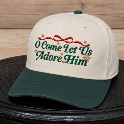 An off-white and dark green baseball cap with embroidered text reading 'O Come Let Us Adore Him' in green, a red bow, and small gold stars, displayed on a black surface against a wood plank background.
