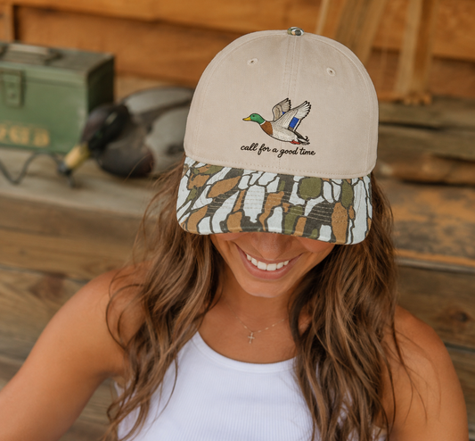 Woman wearing a Call for a Good Time mallard embroidered duck hunting hat with a camouflage brim in a rustic duck camp setting.