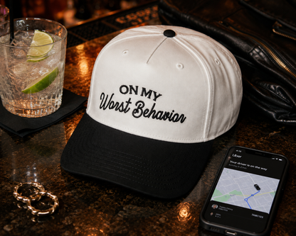 A white and black baseball cap with the embroidered text 'ON MY Worst Behavior' sits on a dark bar counter next to a drink with a lime wedge, gold rings, a black leather jacket, and a smartphone waiting on an Uber ride pickup. 