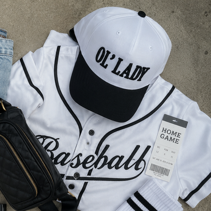 A styled flat-lay of a baseball-themed outfit on a concrete surface, featuring a white and black baseball jersey with 'Baseball' script, a matching cap reading 'OL’ LADY', white socks with black stripes, light-wash jeans, white sneakers, sunglasses, a black quilted crossbody bag, a baseball, and a lip gloss tube.