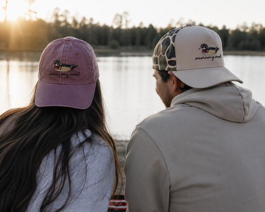 Couple wearing Morning Wood Wood Duck embroidered duck hunting hats while relaxing beside a lake at sunset after a day of waterfowl hunting.