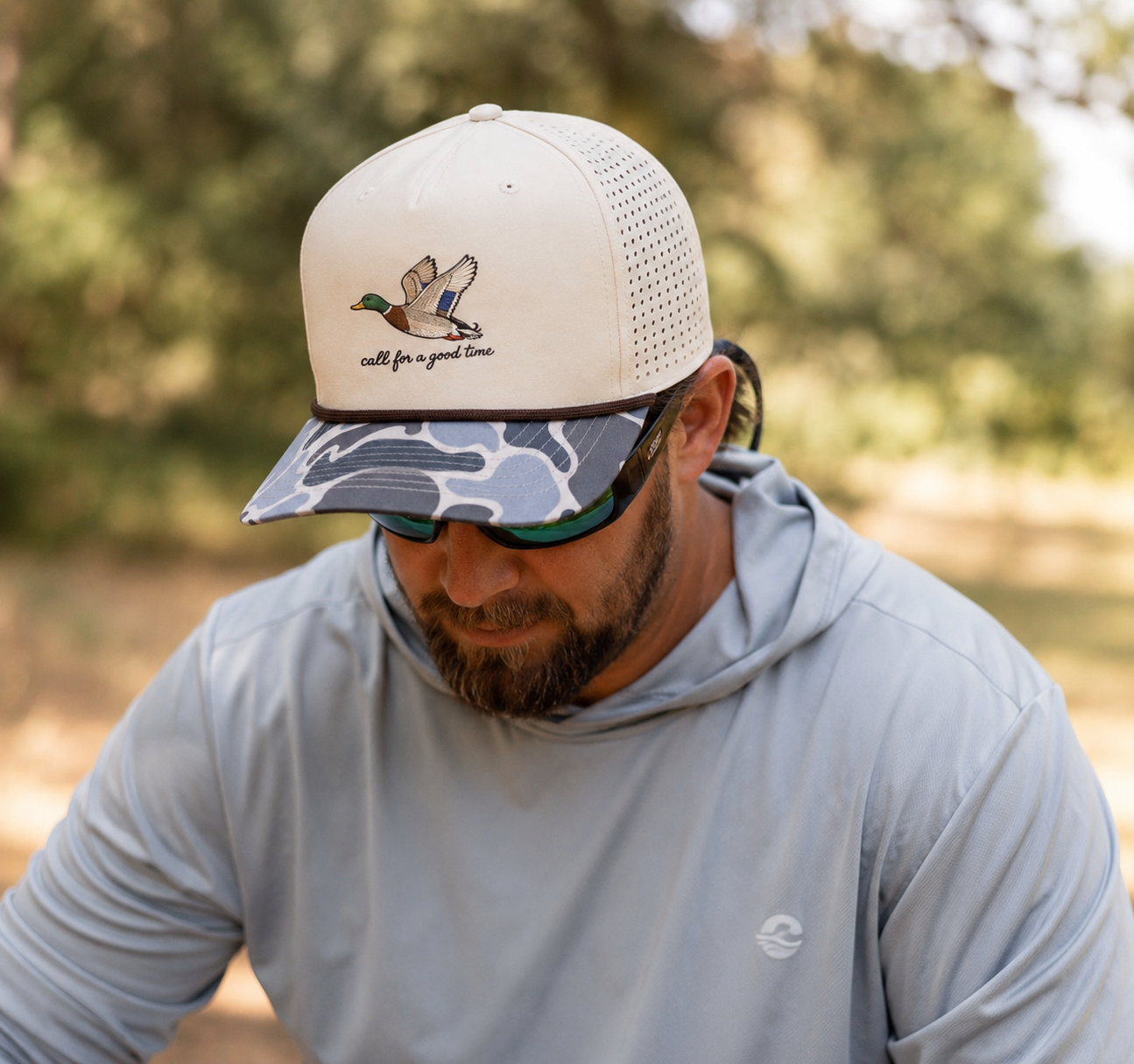 Man wearing a Call for a Good Time mallard embroidered duck hunting performance hat with a perforated back and camouflage brim outdoors.