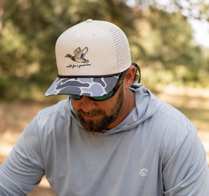 Man wearing a Call for a Good Time mallard embroidered duck hunting performance hat with a perforated back and camouflage brim outdoors.