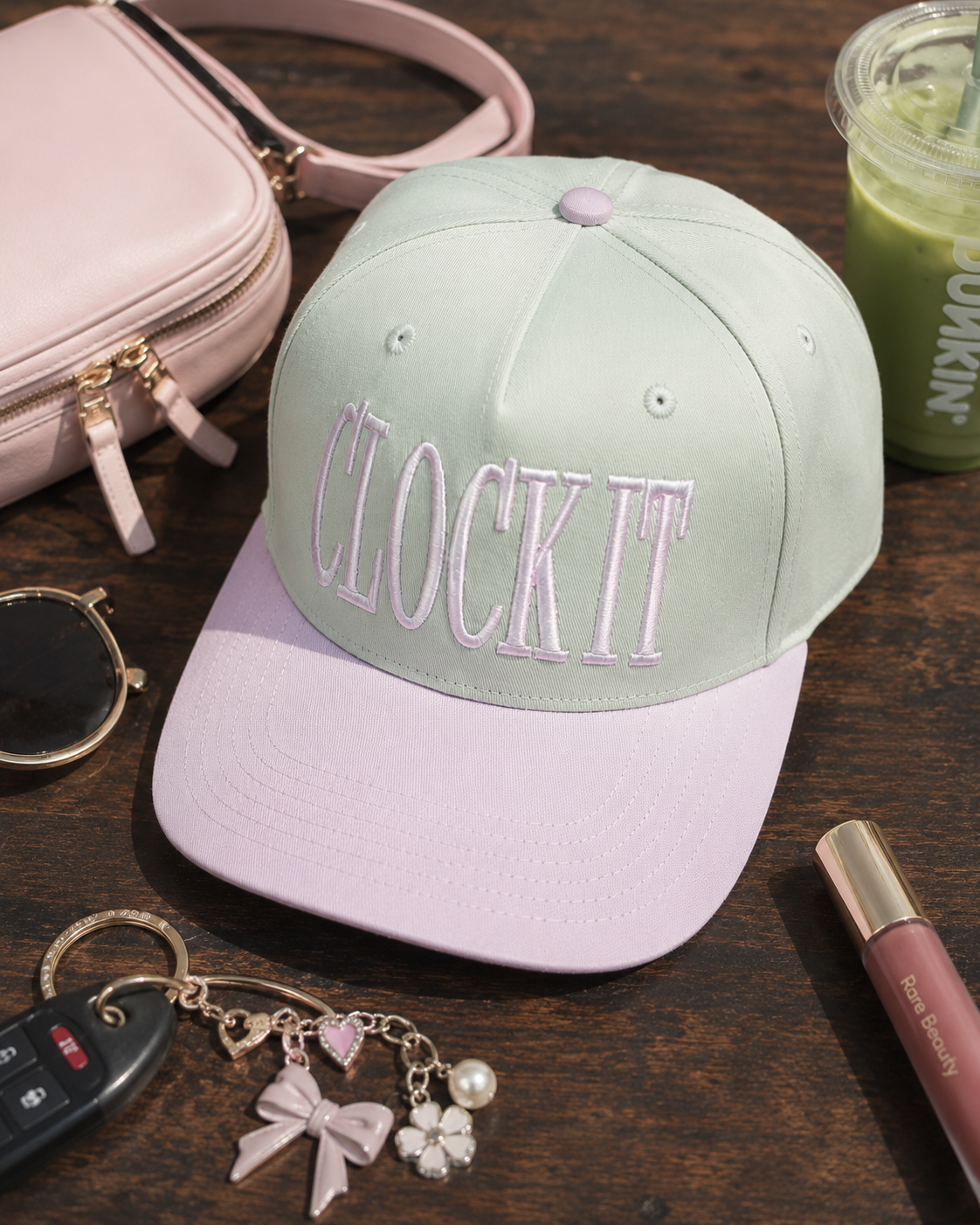 A natural and light pink baseball cap with 'CLOCK IT' embroidered on the front, styled on a dark wooden table with a pink handbag, sunglasses, a keychain, a drink, and a lip gloss nearby.