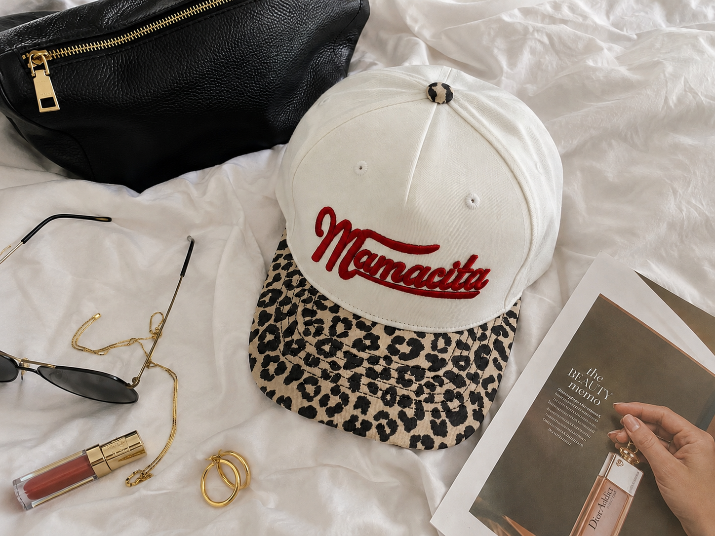 A natural-color baseball cap with a leopard-print visor and red 'Mamacita' embroidery, styled on white bedding with a black pouch, sunglasses, lipstick, rings, and a magazine.