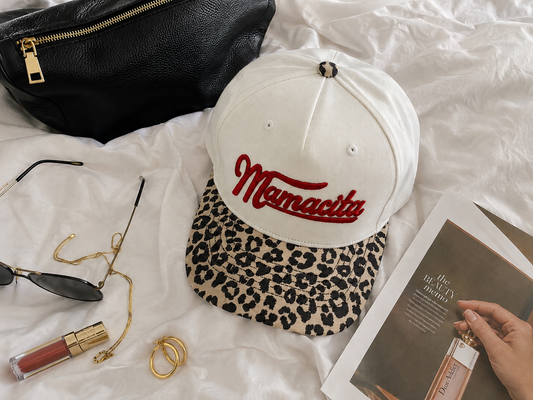 A natural-color baseball cap with a leopard-print visor and red 'Mamacita' embroidery, styled on white bedding with a black pouch, sunglasses, lipstick, rings, and a magazine.
