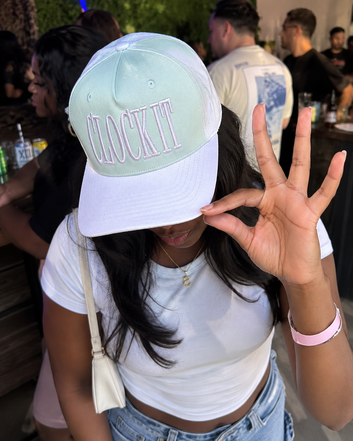A Black girl at a social event wearing a beige and pastel pink baseball cap with the phrase 'CLOCK IT' embroidered in pink on the front, paired with a white crop top and light blue jeans.