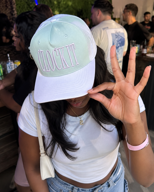 A Black girl at a social event wearing a beige and pastel pink baseball cap with the phrase 'CLOCK IT' embroidered in pink on the front, paired with a white crop top and light blue jeans.
