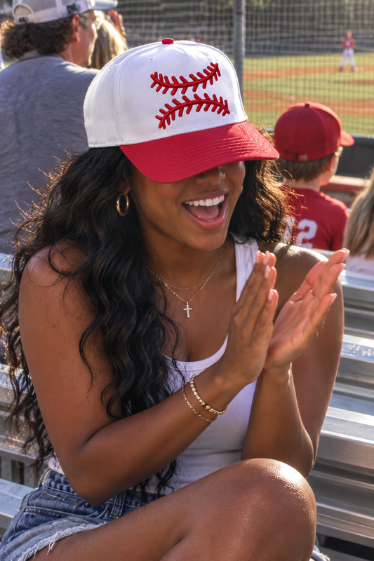 A woman sitting in a baseball field bleachers wearing a white and red baseball cap with red stitching details, clapping her hands in excitement.