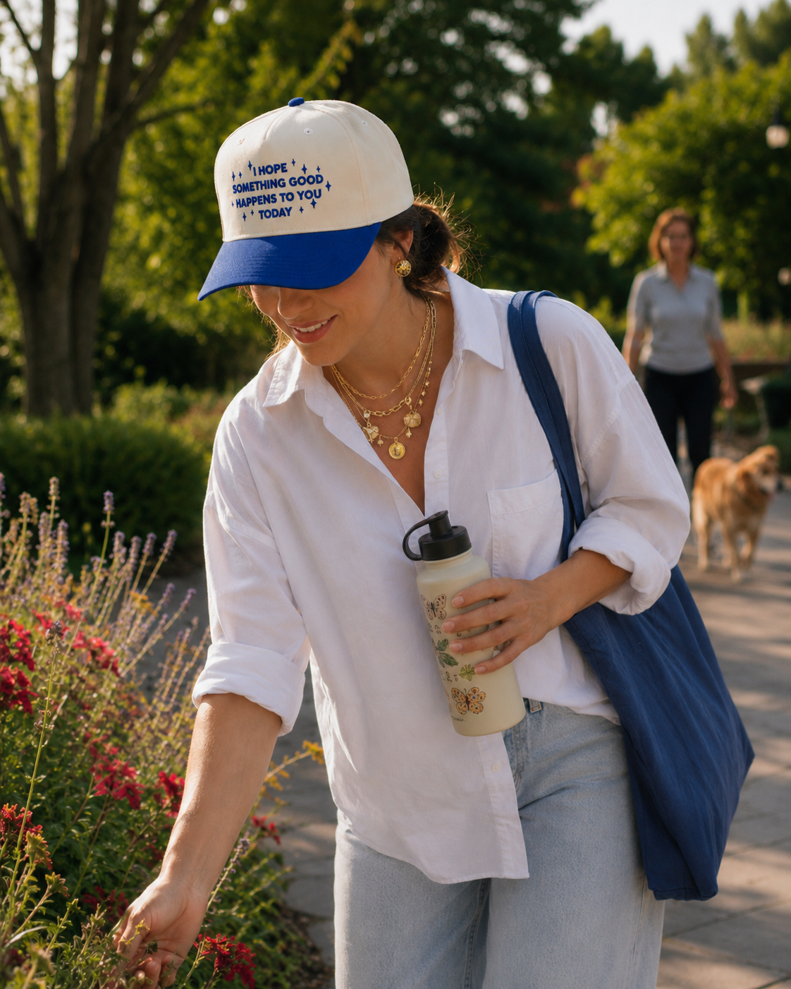 Cream and royal blue embroidered baseball hat with the phrase “I Hope Something Good Happens to You Today” worn by a woman during a morning walk through a botanical garden, lifestyle apparel photography.