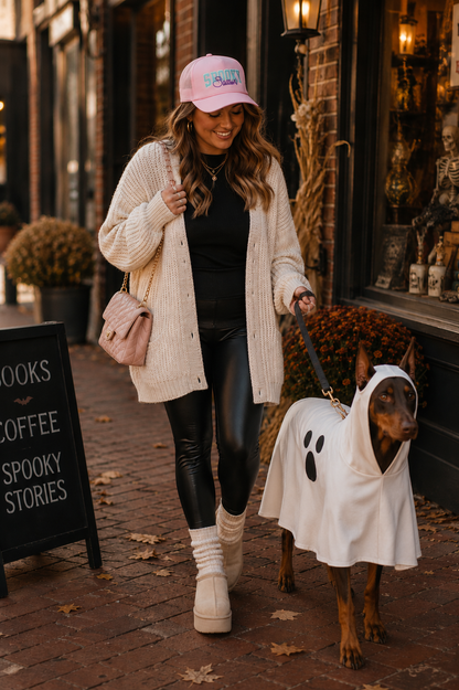 Woman wearing a light pink "Spooky Season" foam trucker hat with mint green chenille embroidery and purple script, walking an adult red Doberman in a ghost costume through a charming historic downtown on a fall afternoon.