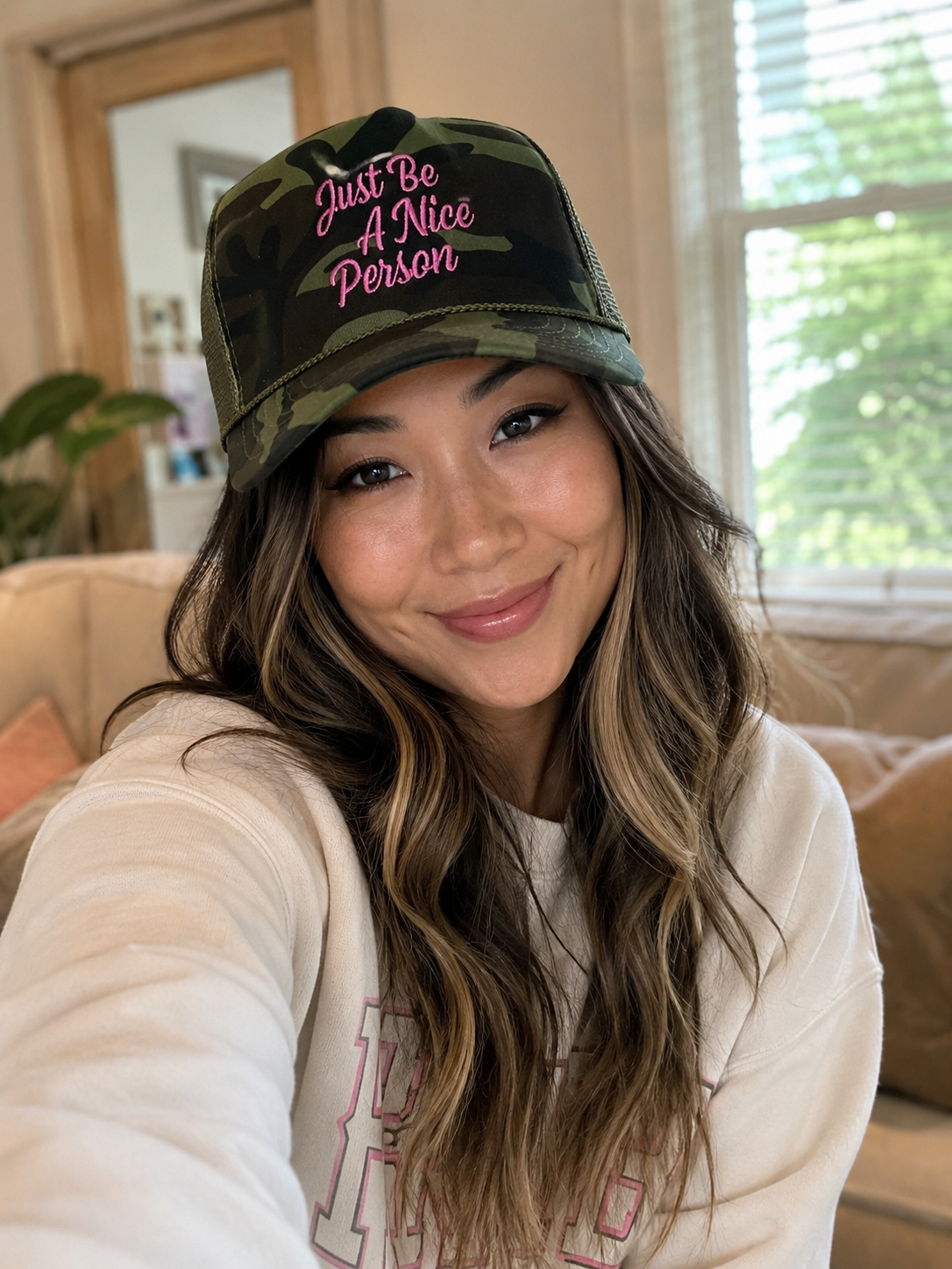 A woman wearing a camouflage trucker hat with pink embroidered text that reads 'Just Be A Nice Person', posing in a cozy indoor setting.