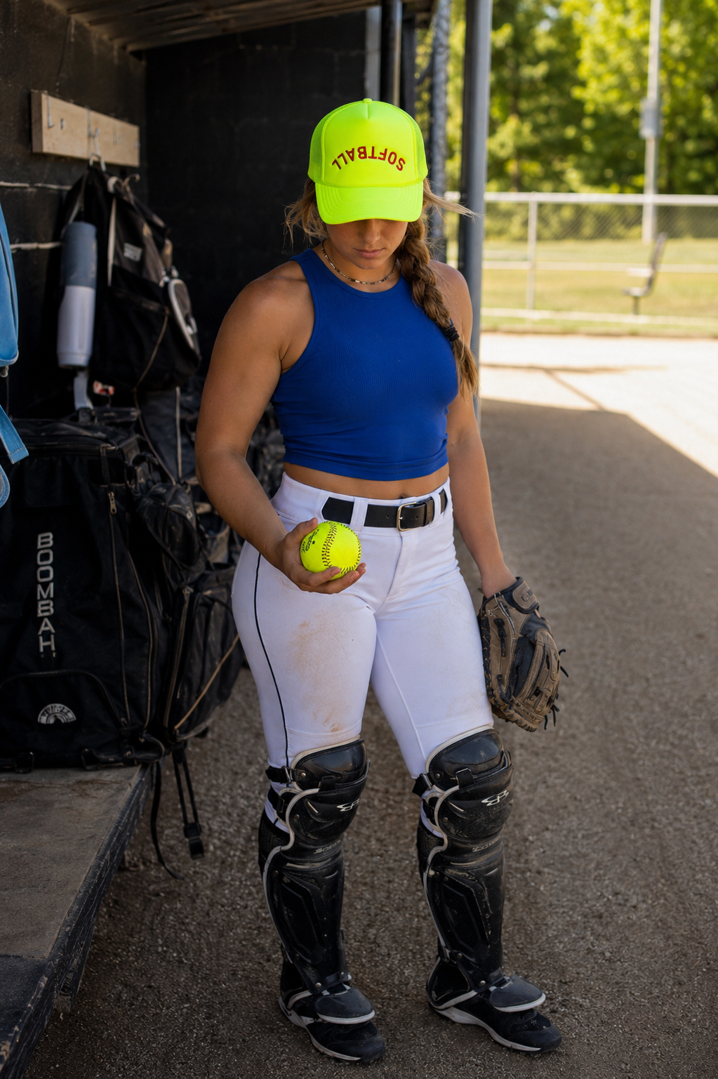 A softball player stands in a dugout holding a yellow ball, wearing a neon yellow cap with 'SOFTBALL' in red, a blue tank top, white pants, black shin guards, and a glove.