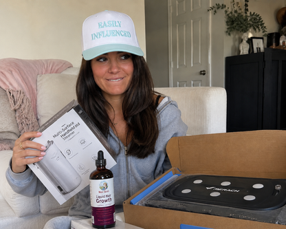 A woman wearing a natural and light blue 'EASILY INFLUENCED' trucker hat sits in a living room setting, holding a product box and a dropper bottle labeled 'Liquid Root Growth'. An open cardboard box containing a black vibration plate sits on a table in the foreground.