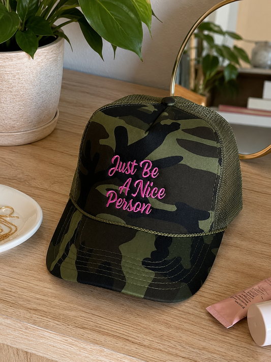 A camouflage trucker hat with pink embroidered text reading 'Just Be A Nice Person' sits on a wooden table in a styled indoor setting with a potted plant and mirror in the background.