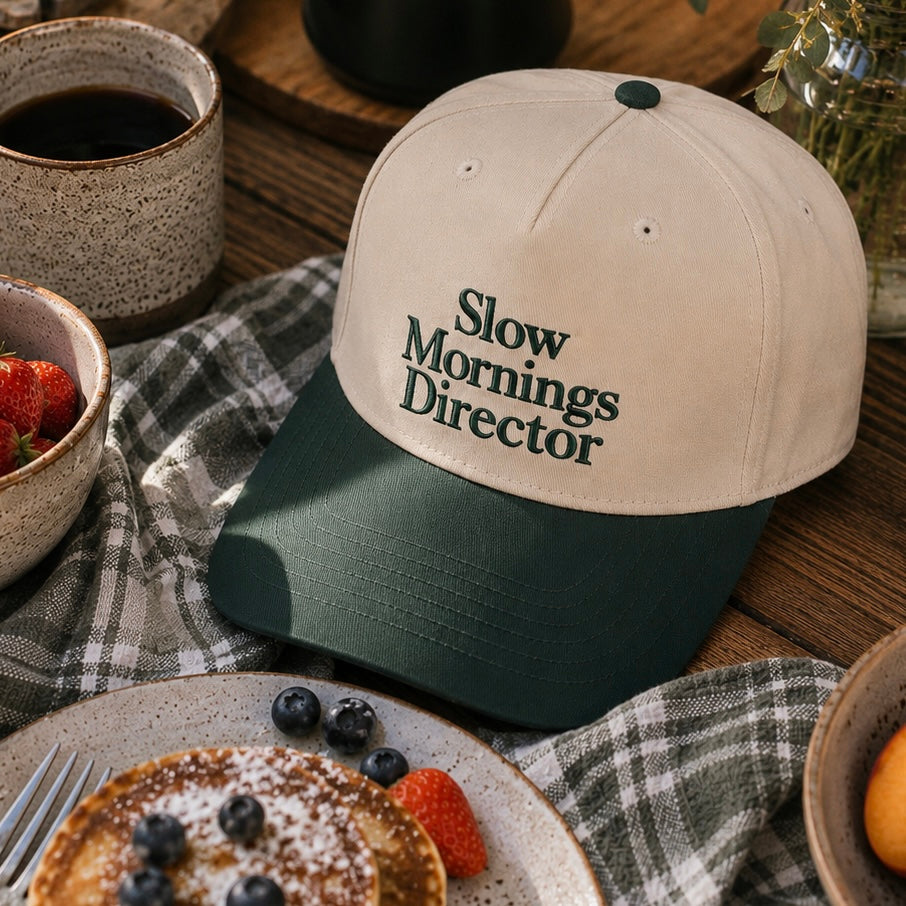 A two-tone baseball cap with a cream crown and a dark forest green brim, embroidered with the text 'Slow Mornings Director' in dark green. The cap is styled in a cozy breakfast setting on a plaid napkin with pancakes, blueberries, strawberries, and a cup of coffee.