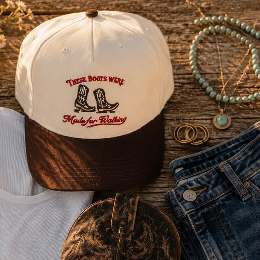A cream and brown baseball cap with embroidered text reading 'These Boots Were Made for Walking' and a cowboy boots graphic, styled on a rustic wood surface with jeans, jewelry, and a white shirt nearby.