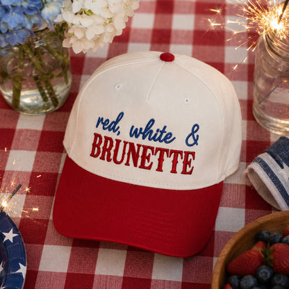 A baseball cap with a natural crown and red visor, embroidered with 'red, white & BRUNETTE' in blue and red lettering, styled on a red and white checkered tablecloth with flowers, sparklers, and a bowl of berries.