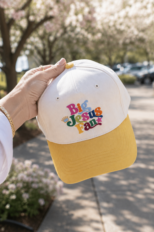 A hand holds a baseball cap outdoors with a blurred background of blooming trees and a sidewalk. The cap has a beige crown and a mustard yellow brim, with colorful embroidered text reading 'Big Jesus Fan' on the front.