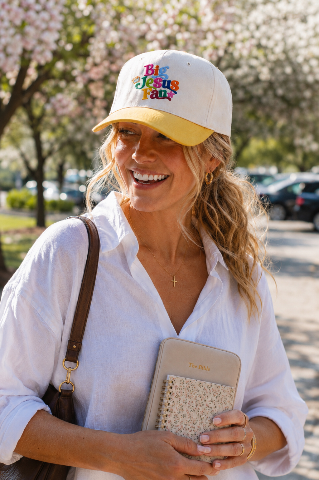 A woman outdoors wearing a natural baseball cap with a yellow brim and colorful 'Big Jesus Fan' text, holding a beige clutch. She is wearing a white button-down shirt and a brown shoulder bag.