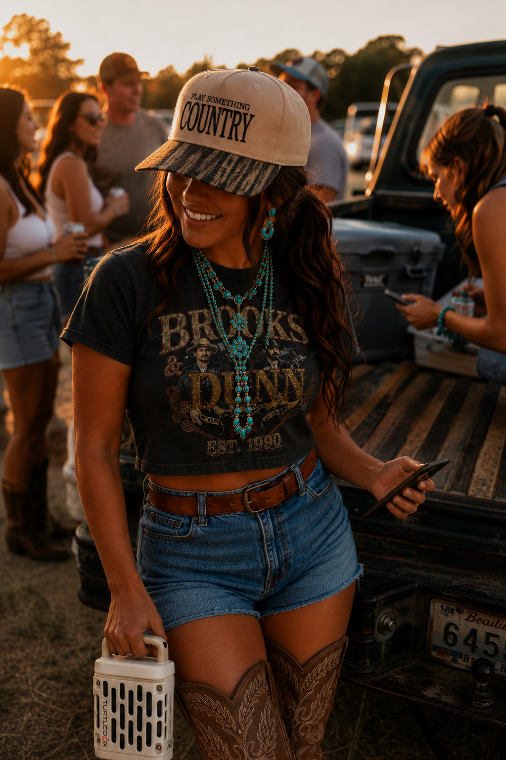 A woman outdoors at sunset wearing a beige baseball cap with 'PLAY SOMETHING COUNTRY' printed on it, a black graphic T-shirt, denim shorts, and cowboy boots, standing near the back of a pickup truck.