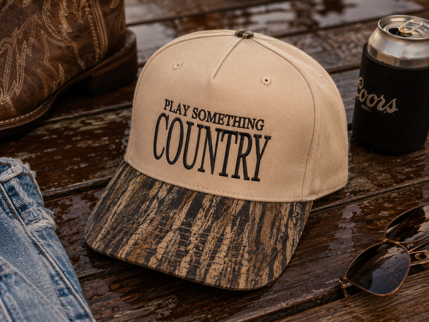 A beige baseball cap with black text reading 'PLAY SOMETHING COUNTRY' on the front, paired with a brown camouflage-patterned brim, styled on a wet wooden surface with a boot, jeans, sunglasses, and a Coors can nearby.