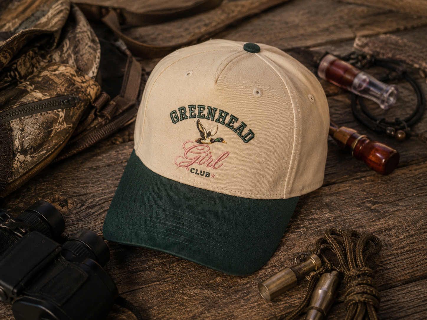 A baseball cap with a cream crown and dark green visor, featuring 'GREENHEAD Girl CLUB' embroidered on the front, styled on a rustic wooden surface with hunting accessories.