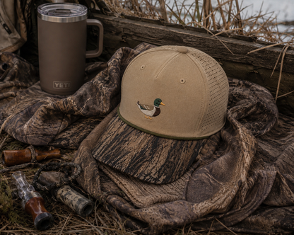 A tan mesh-back baseball cap with a small mallard duck embroidery on the front, styled on camouflage fabric next to a YETI tumbler and duck hunting props.