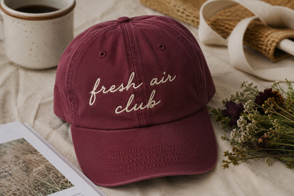 A burgundy dad hat baseball cap with 'fresh air club' embroidered in beige script on the front, styled on a beige fabric surface with a ceramic mug, dried flowers, and a notebook.